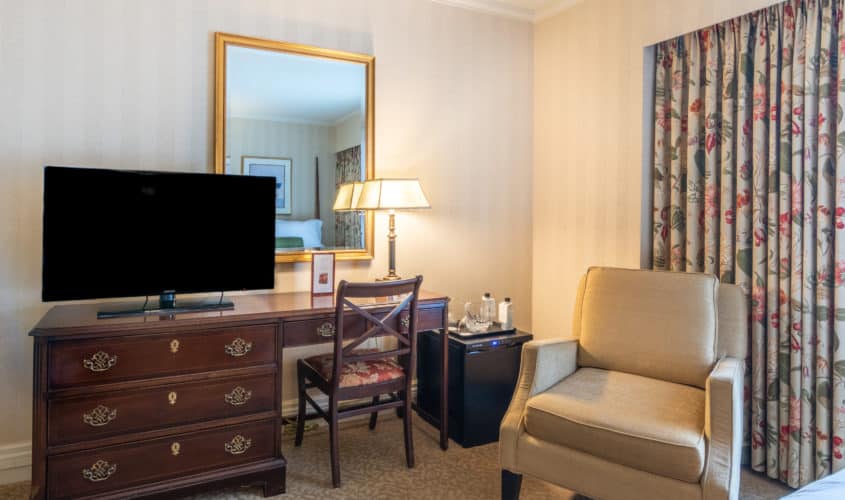 Our Seattle deluxe queen accommodations featuring a desk with small fridge & large mirror reflecting the guest bed and window.