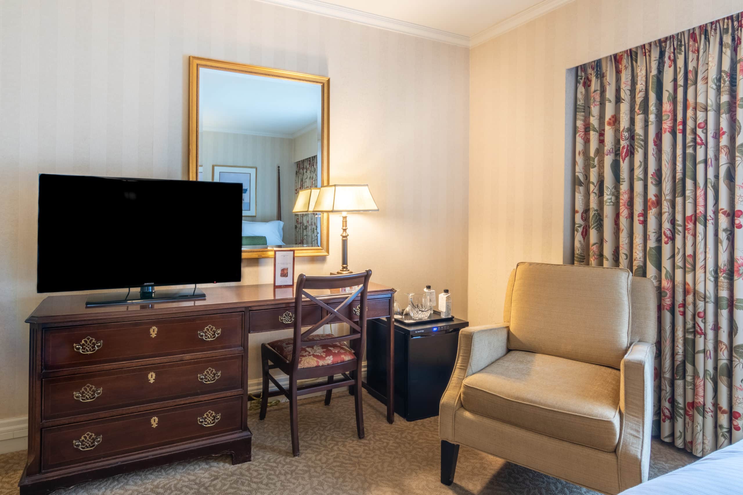 Our Seattle deluxe queen accommodations featuring a desk with small fridge & large mirror reflecting the guest bed and window.