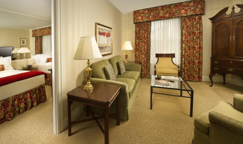 Seattle hotel Deluxe Queen Suite Parlor with green sofa, glass coffee table and red drapes with a view of the Bedroom with 2 beds.