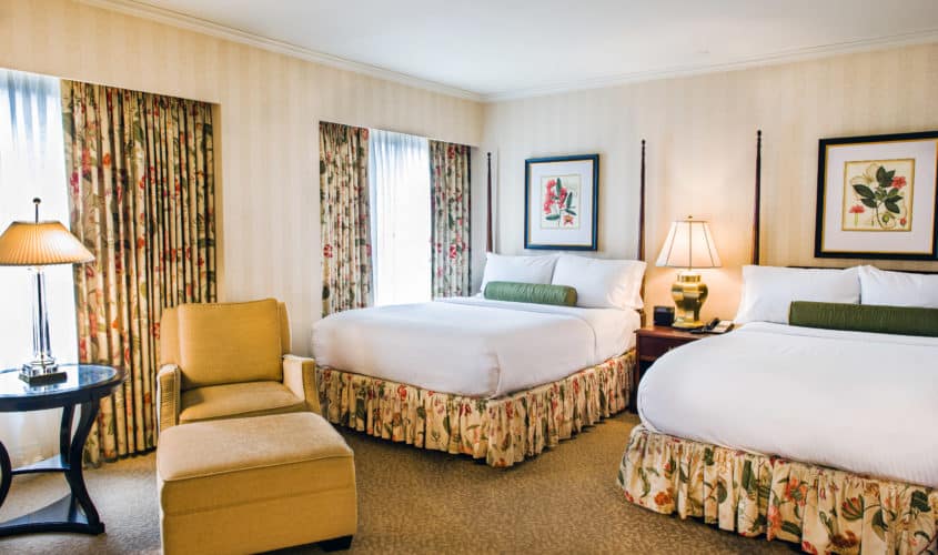 The Mayflower Hotel's Classic Double Queen Guestroom with floral patterns, large window, high ceilings and chair.