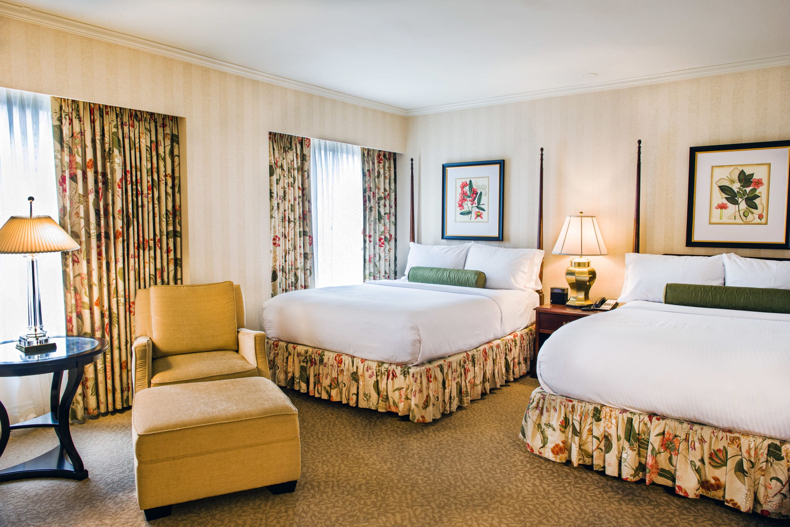 The Mayflower Hotel's Classic Double Queen Guestroom with floral patterns, large window, high ceilings and chair.